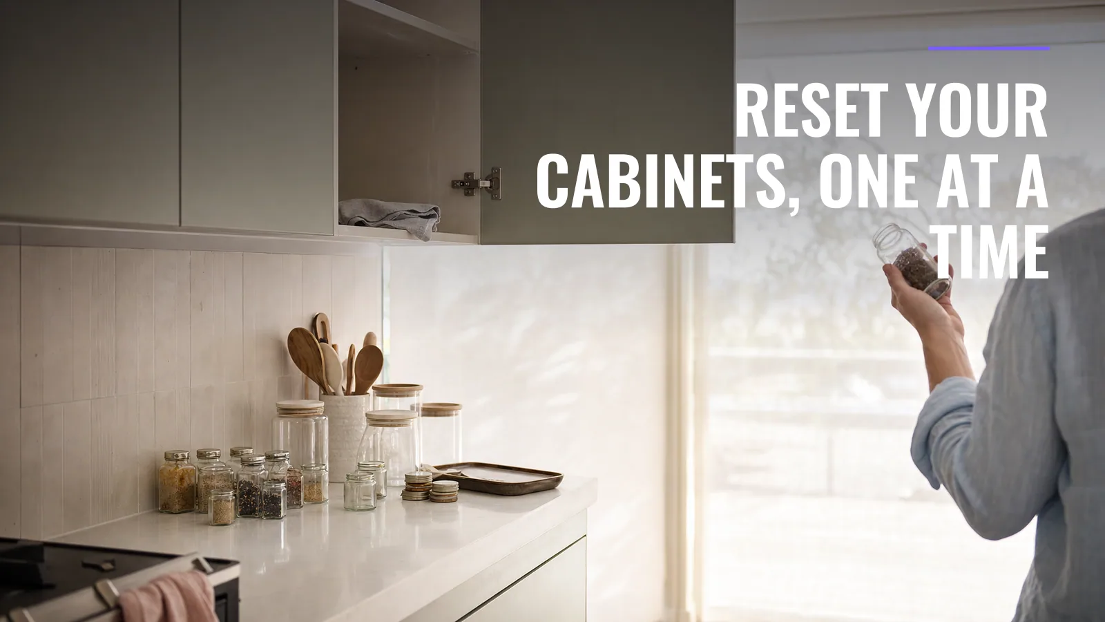 An open, emptied kitchen wall cabinet with a bare wiped shelf, its contents spread along the stone benchtop below, and a forearm holding up a spice jar.